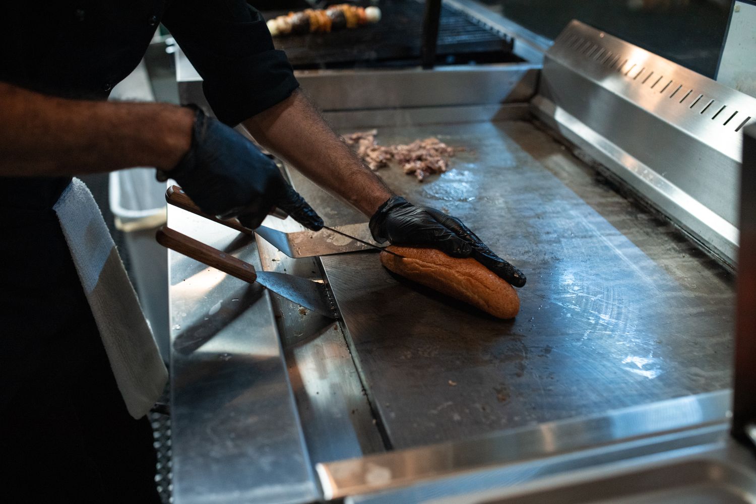 A cook working a roll on the flat-top griddle beside chopped grilled meat