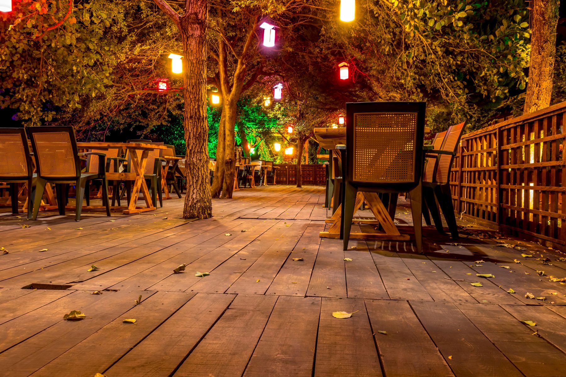 An outdoor deck set with tables and chairs under warm hanging lights in the evening